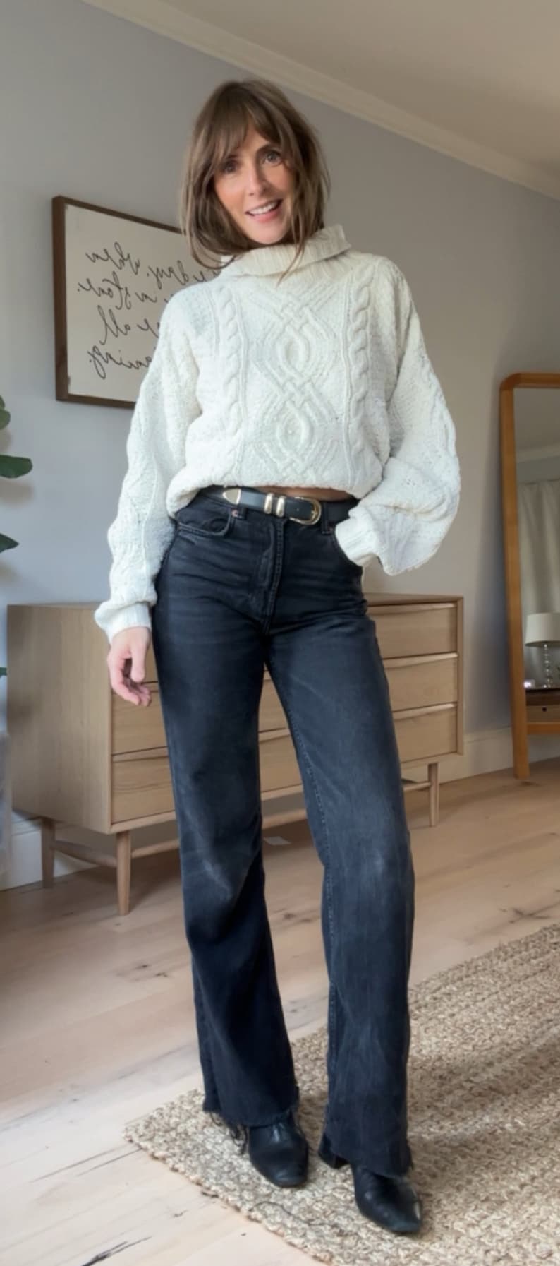 May include: A woman wearing a white cable knit jumper with a high neck and black jeans. She is wearing a silver belt with a buckle.