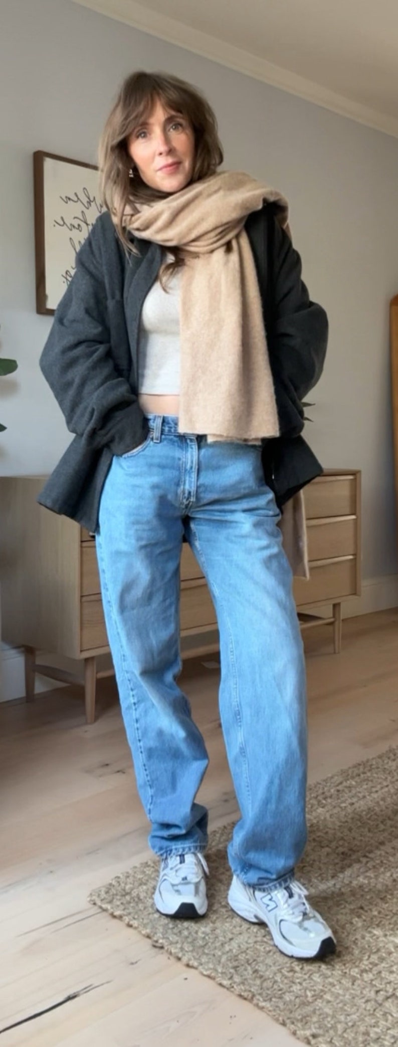 May include: A woman wearing a dark grey jacket with a light brown scarf, light blue jeans, and white trainers.