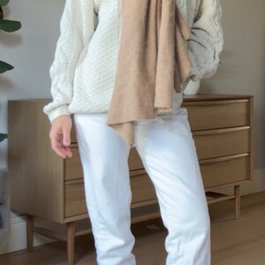 May include: A woman wearing a white cable knit jumper with a brown scarf, white jeans, and white trainers.