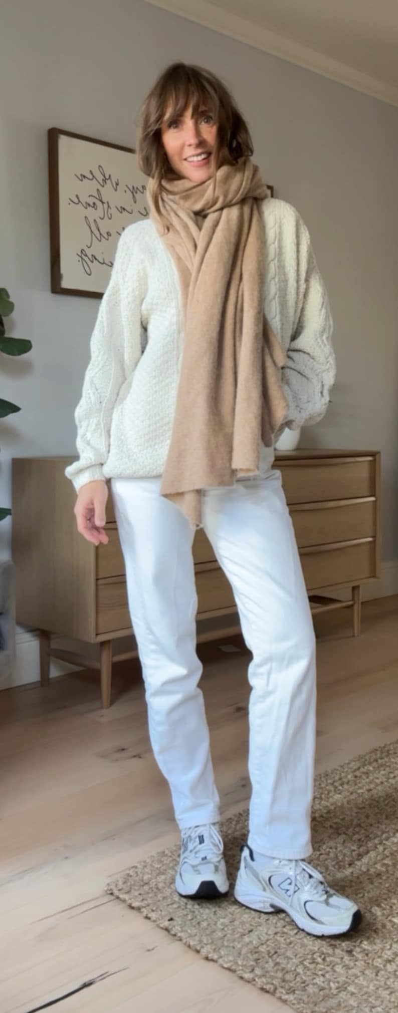 May include: A woman wearing a white cable knit jumper with a brown scarf, white jeans, and white trainers.