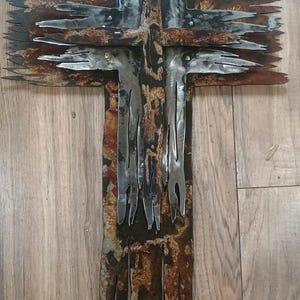 May include: A metal cross with a distressed, weathered appearance. The cross features a layered design with jagged edges and a mix of silver and rust-coloured metal. The artwork is mounted on a wooden surface.