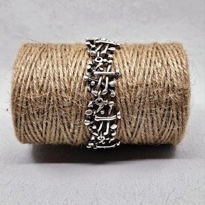 May include: A silver-colored bracelet with an abstract design is displayed on a spool of brown twine. The bracelet features a series of interconnected shapes and lines. The twine is tightly wound, creating a textured background.