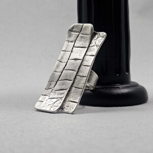 May include: A silver ring with a textured, rectangular design. The ring is made of metal and has a unique, geometric shape.
