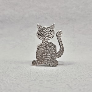 May include: A silver cat-shaped ring with a textured finish. The ring depicts a cat silhouette with a raised tail and pointed ears. A unique piece of jewellery.