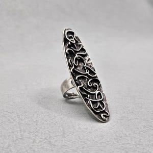 May include: A silver-toned ring with an elongated oval face. The face features an intricate, swirling design. The ring's band is simple and connects to the back of the oval. The ring is set against a gray background.