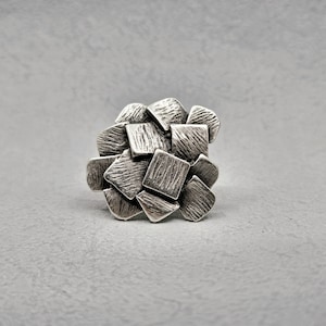 Può includere: Un anello d'argento con un design a grappolo. L'anello presenta molteplici forme strutturate, quadrate e semicerchi, disposte insieme. Il metallo ha un aspetto leggermente appannato, che gli conferisce un aspetto vintage. L'anello è posto su uno sfondo grigio.