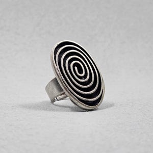 Puede incluir: Anillo plateado con una cara ovalada con un diseño de espiral negro. El anillo tiene una banda ancha y un acabado texturizado y envejecido. El diseño en espiral es un elemento audaz y llamativo.
