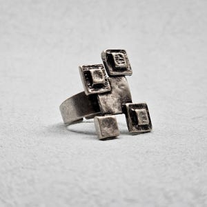 May include: A silver ring with a geometric design. The ring features five square-shaped pieces, each with a raised center. The ring is made of metal and has a textured finish.