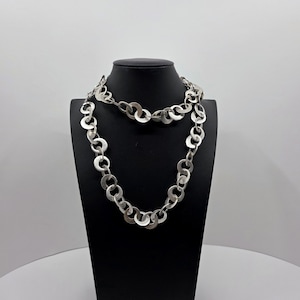 May include: A silver-toned necklace with a series of connected, open circle links. The necklace is draped over a black mannequin bust, showcasing its design. The background is a plain white, highlighting the jewelry's details.
