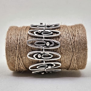 Puede incluir: Pulsera de plata con un diseño geométrico de círculos y óvalos entrelazados. La pulsera está sobre un carrete de cuerda marrón.