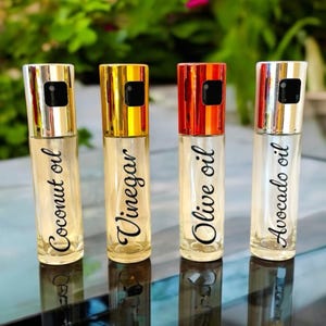 May include: Four clear glass spray bottles with silver, gold, red, and silver caps. The bottles are labeled with black text: "Coconut oil", "Vinegar", "Olive oil", and "Avocado oil".