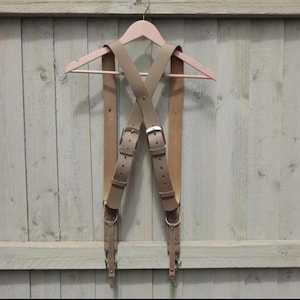 May include: Tan leather camera straps with metal buckles and clips, hanging on a wooden hanger against a light wood background.