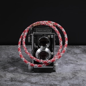 May include: A classic Mamiyaflex camera with a red, white, and grey patterned camera strap. The black camera has two lenses and the text "MAMIYAFLEX". The strap is looped around the camera body.