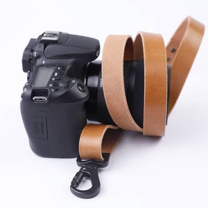 Może przedstawiać: Czarny aparat DSLR z brązowym skórzanym paskiem do aparatu. Pasek jest przymocowany do aparatu za pomocą czarnego metalowego zatrzasku. Obiektyw aparatu jest czarny z nadrukiem "SIGMA".