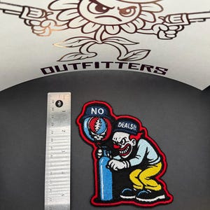 Op de afbeelding: Geborduurde patch met een clown met een rode neus en een sinistere grijns, die een blauwe tank vasthoudt. De patch heeft de tekst "NO DEALS!" en is rood omlijnd. Een liniaal staat naast de patch. De achtergrond heeft een bloem met pistolen.