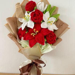 May include: A handcrafted bouquet of crocheted flowers, featuring red roses, white lilies, and red poinsettias. The bouquet is wrapped in brown paper and tied with a brown and cream ribbon. Green leaves and yellow stamens add detail.