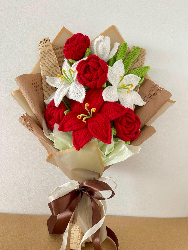 May include: A handcrafted bouquet of crocheted flowers, featuring red roses, white lilies, and red poinsettias. The bouquet is wrapped in brown paper and tied with a brown and cream ribbon. Green leaves and yellow stamens add detail.