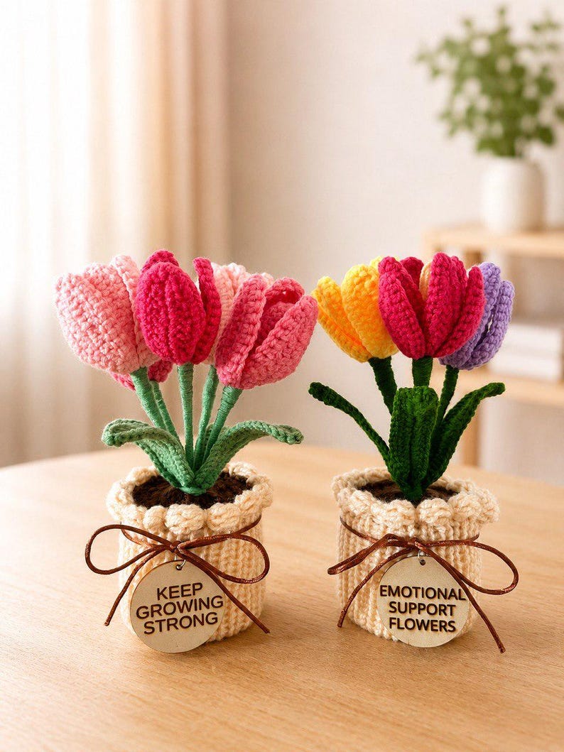 Może przedstawiać: Dwie szydełkowane kompozycje tulipan&oacute;w w beżowych doniczkach. Lewa doniczka ma drewnianą zawieszkę z napisem KEEP GROWING STRONG. Prawa ma napis EMOTIONAL SUPPORT FLOWERS. Tulipany są r&oacute;żowe, ż&oacute;łte i fioletowe z zielonymi łodygami oraz liśćmi.