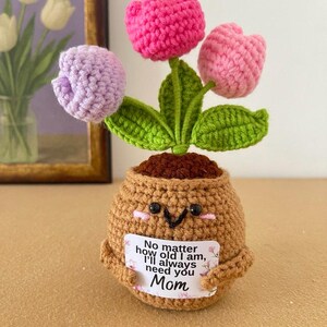 May include: A crocheted potted plant featuring three tulip flowers in shades of pink, light pink, and lavender with green leaves. The brown pot has a smiling face and holds a white sign that reads, No matter how old I am, I'll always need you Mom.