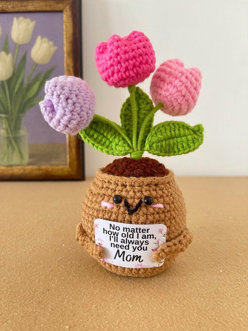 May include: A crocheted potted plant featuring three tulip flowers in shades of pink, light pink, and lavender with green leaves. The brown pot has a smiling face and holds a white sign that reads, No matter how old I am, I'll always need you Mom.
