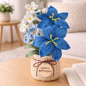 Op de afbeelding: Een gehaakt bloemstuk in een beige pot, met grote blauwe bloemen met gele accenten en kleinere witte en blauwe bloemen. Een houten label met "HAPPY BIRTHDAY" is bevestigd. Het arrangement staat op een houten tafel.