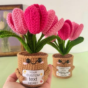 May include: Two crocheted potted tulips with pink petals and green leaves. The brown pots feature embroidered faces with black eyes and pink cheeks. Each pot holds a white rectangular sign; one reads "YOUR CUSTOM text HERE!" and the other "THANK YOU FOR helping me GROW".