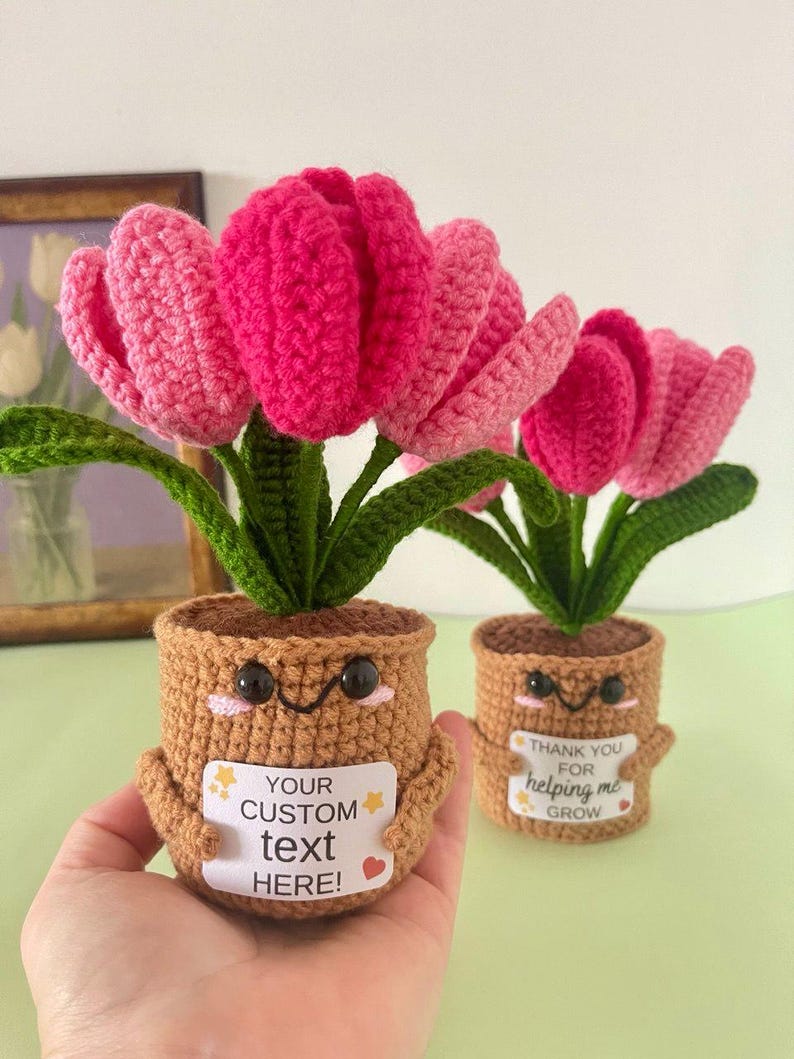May include: Two crocheted potted tulips with pink petals and green leaves. The brown pots feature embroidered faces with black eyes and pink cheeks. Each pot holds a white rectangular sign; one reads "YOUR CUSTOM text HERE!" and the other "THANK YOU FOR helping me GROW".