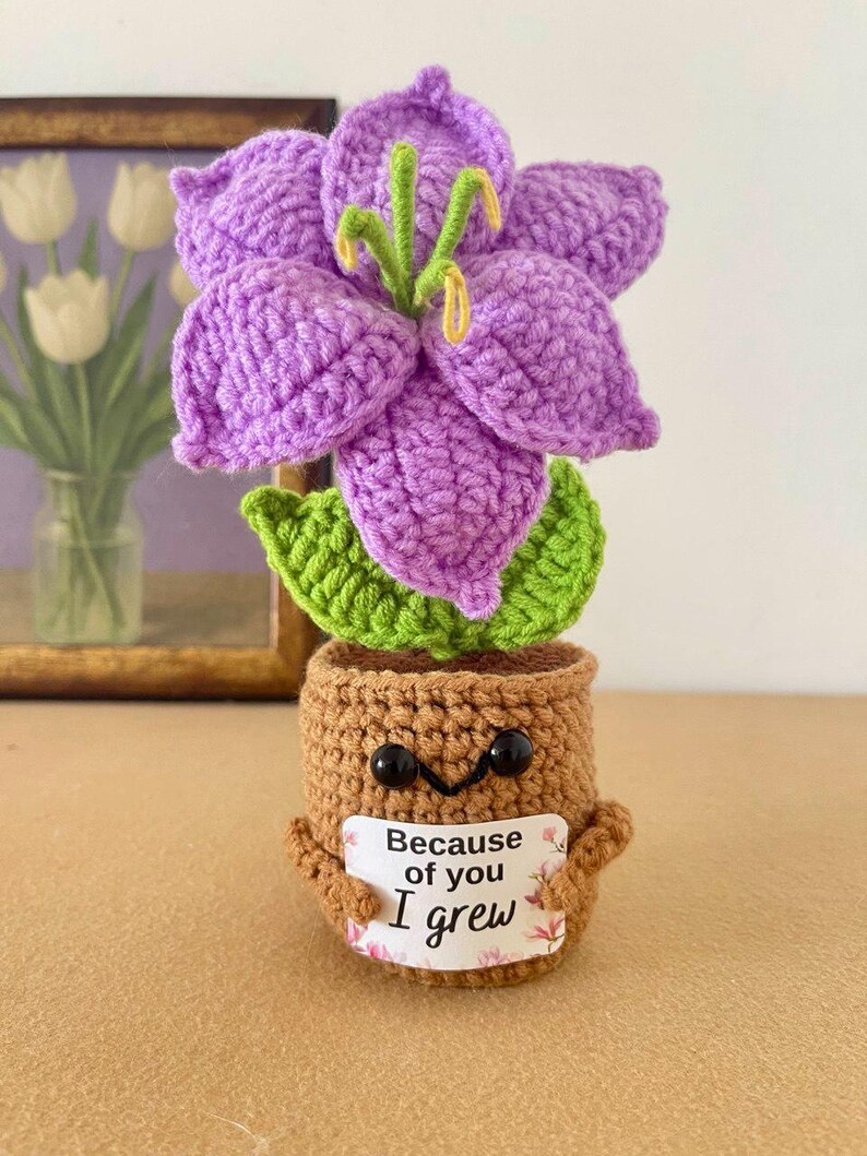 May include: A crocheted purple lily flower with green leaves in a brown pot. The pot has two black bead eyes and a small sign held by two arms that reads, "Because of you I grew," with a floral border. The flower has yellow stamens in the center.