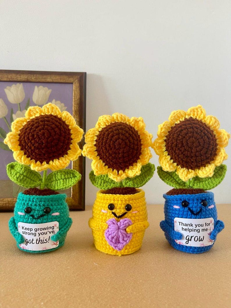 May include: Three crocheted sunflower plants in colorful pots. The teal pot says "Keep growing strong you've got this!", the yellow pot holds a purple heart, and the blue pot says "Thank you for helping me grow". Each has black eyes and pink cheeks.