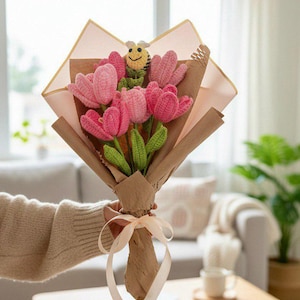 May include: A bouquet of pink crochet tulips with green leaves and a yellow and black crochet bee. The flowers are wrapped in brown paper and tied with a cream ribbon. The bouquet is held by a person.