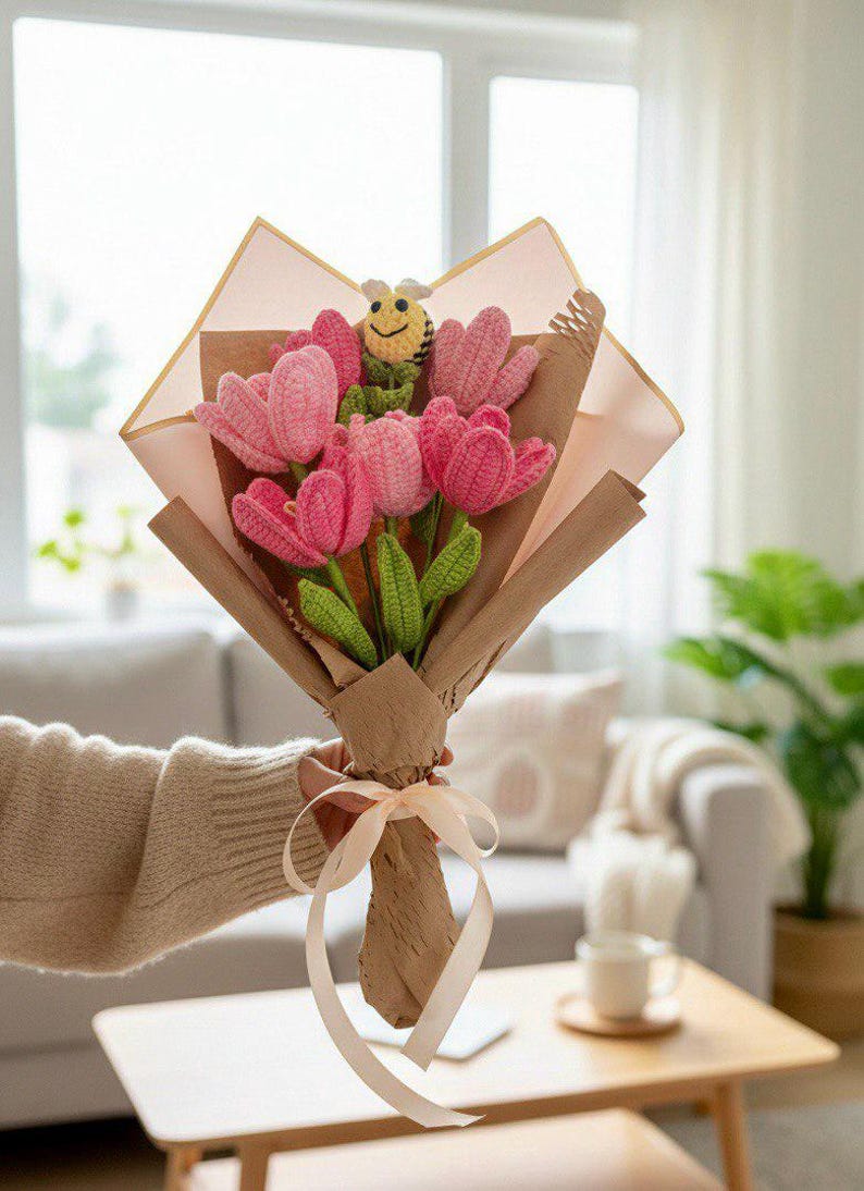 May include: A bouquet of pink crochet tulips with green leaves and a yellow and black crochet bee. The flowers are wrapped in brown paper and tied with a cream ribbon. The bouquet is held by a person.