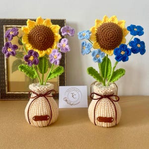 Könnte beinhalten: Zwei gehäkelte Blumenarrangements in beigen Strickvasen. Die linke Vase enthält eine gelbe Sonnenblume und violette Blumen. Die rechte Vase enthält eine gelbe Sonnenblume und blaue Blumen. Jede Vase hat ein braunes Band und ein Etikett mit Hand made.