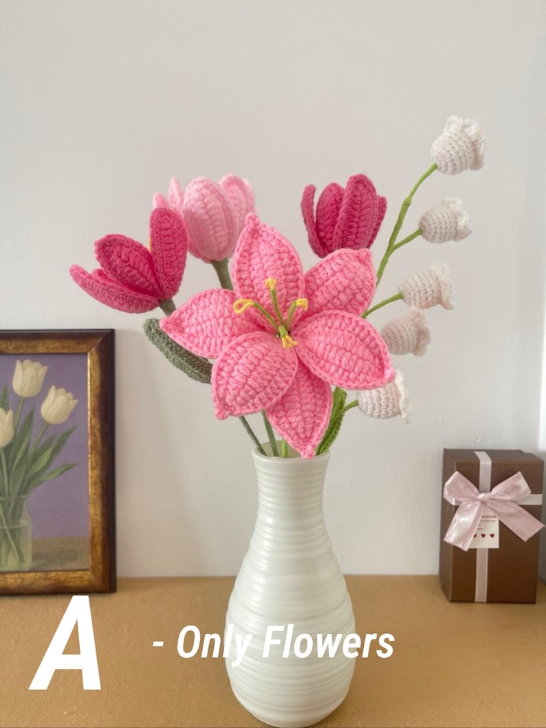 May include: A white ceramic vase holds a bouquet of handmade crochet flowers. The bouquet includes pink lilies, tulips, and white bell-shaped flowers. The text "A - Only Flowers" is visible in the image.