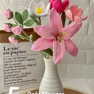 May include: A bouquet of handmade crochet flowers in a white vase. The arrangement includes pink lilies, white and pink tulips, and pink buds with green leaves. The vase sits in front of a paper bag with French text.
