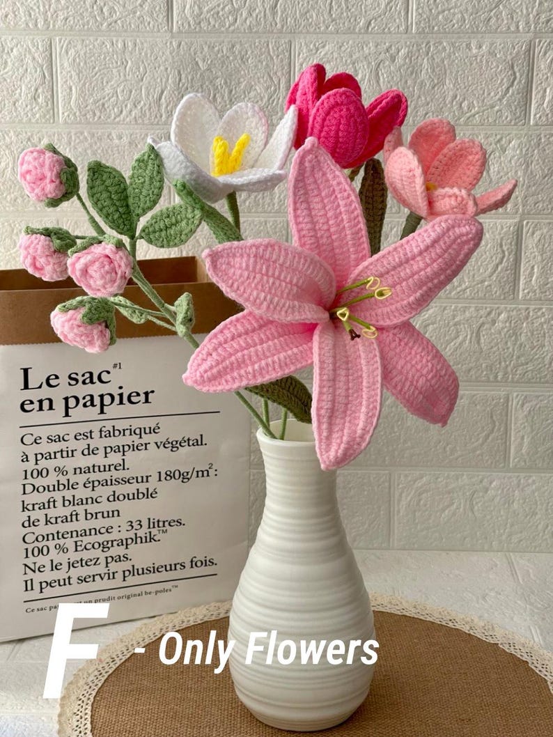 May include: A bouquet of handmade crochet flowers in a white vase. The arrangement includes pink lilies, white and pink tulips, and pink buds with green leaves. The vase sits in front of a paper bag with French text.