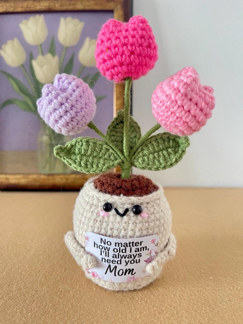 May include: A crocheted potted plant featuring three flowers in shades of hot pink, light pink, and lavender with green leaves. The beige pot has a smiling face and holds a white sign that reads, No matter how old I am, I'll always need you Mom.