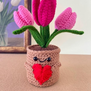 May include: A crocheted plant in a tan pot with a smiling face and a red heart attached to the front. The plant features three tulips in shades of bright pink and light pink, with green stems and leaves extending upwards from the top of the pot.