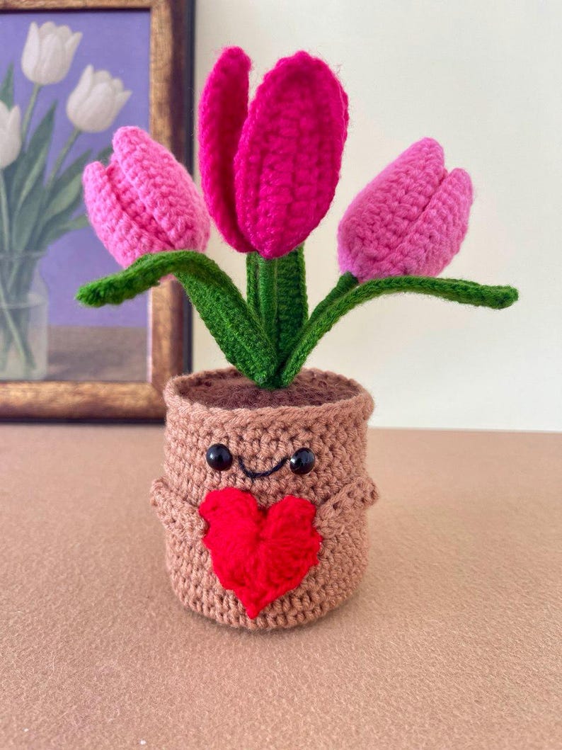 May include: A crocheted plant in a tan pot with a smiling face and a red heart attached to the front. The plant features three tulips in shades of bright pink and light pink, with green stems and leaves extending upwards from the top of the pot.