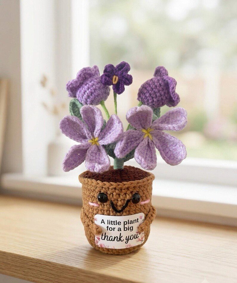 May include: A crocheted potted plant with purple flowers and green stems in a brown textured pot. The pot features a smiling face with black eyes and pink cheeks, holding a white sign that reads, A little plant for a big thank you.