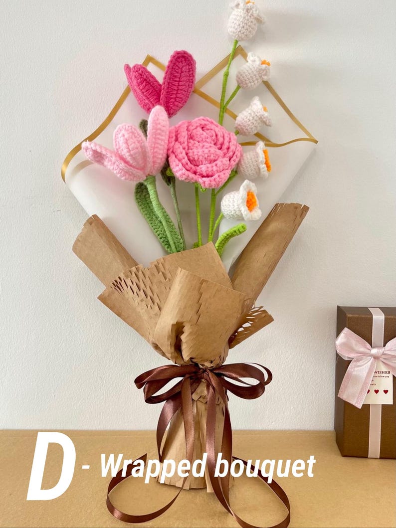 May include: A handmade crochet bouquet featuring pink and white flowers with green stems, wrapped in brown paper and tied with a brown ribbon. The bouquet includes tulips, roses, and other floral designs. The text "D - Wrapped bouquet" is visible.