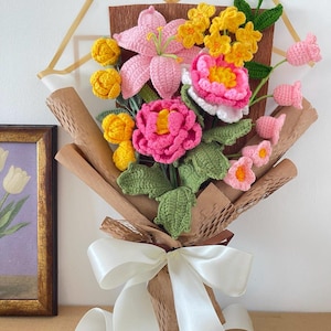 Op de afbeelding: Een handgemaakt gehaakt boeket met roze lelies, gele en roze rozen en andere kleurrijke bloemen. Het boeket is verpakt in bruin papier en vastgebonden met een wit satijnen lint. Groene bladeren geven het arrangement een natuurlijke uitstraling.