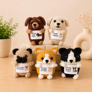 Könnte beinhalten: Fünf kleine gehäkelte Hundefiguren in verschiedenen Farben und Rassen, die jeweils ein weißes rechteckiges Schild mit Text und einer passenden Illustration halten. Die Hunde umfassen einen braunen Dackel, einen beigen Hund, einen hellbraunen Mops, einen gelben Corgi sowie einen schwarz-weißen Hund.