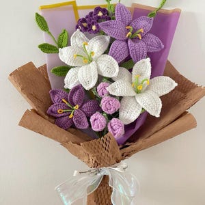 May include: A handmade crochet bouquet featuring white and purple lilies, small pink buds, and green leaves. The flowers are wrapped in brown paper and purple tissue, tied with an iridescent ribbon.