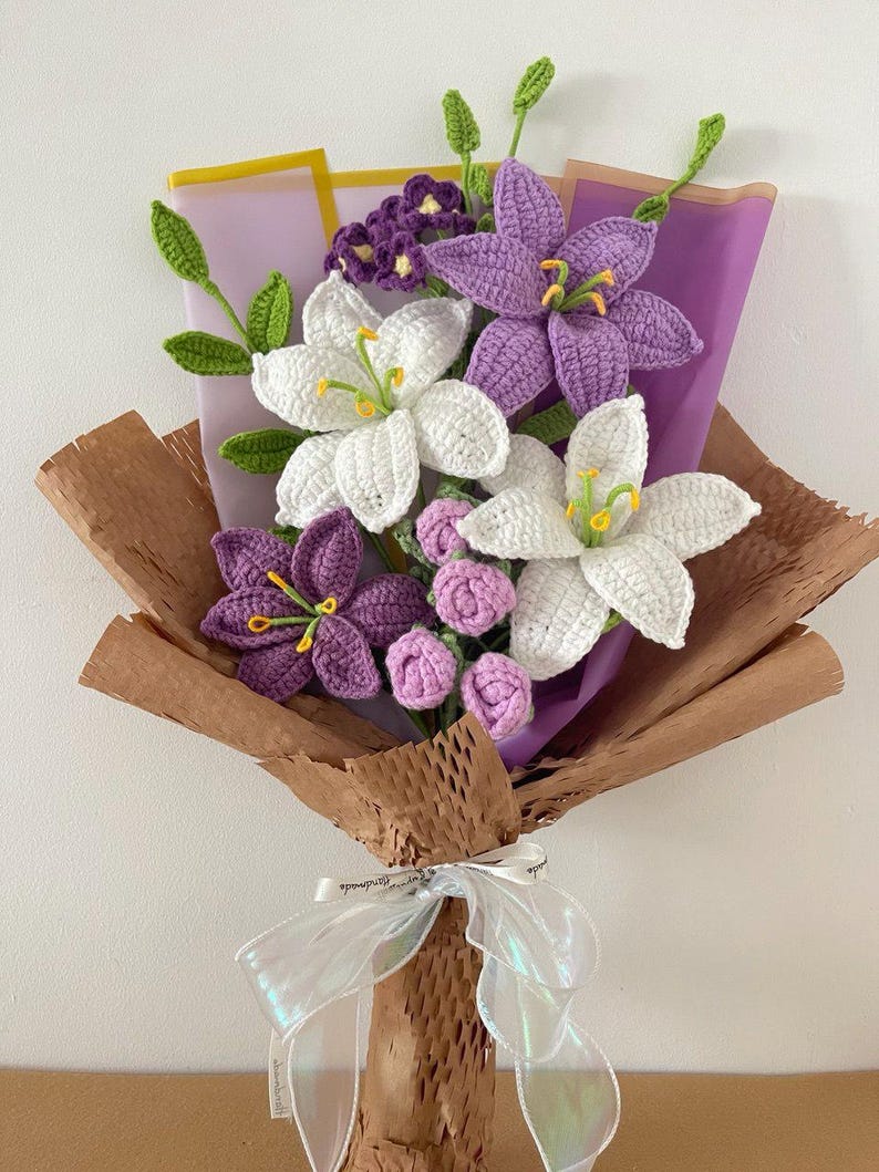 May include: A handmade crochet bouquet featuring white and purple lilies, small pink buds, and green leaves. The flowers are wrapped in brown paper and purple tissue, tied with an iridescent ribbon.