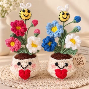 May include: Two crocheted potted plants with smiling faces and red hearts on the pots. Each pot contains green stems with colourful flowers and a small bee on top. One pot has a wooden tag that reads LOVE YOU MOM in black text.