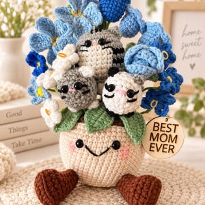 May include: A crocheted arrangement featuring a cream-colored pot with a smiling face and brown feet, holding three small crocheted animals and blue and white flowers. A round wooden tag attached to the side displays the text BEST MOM EVER.