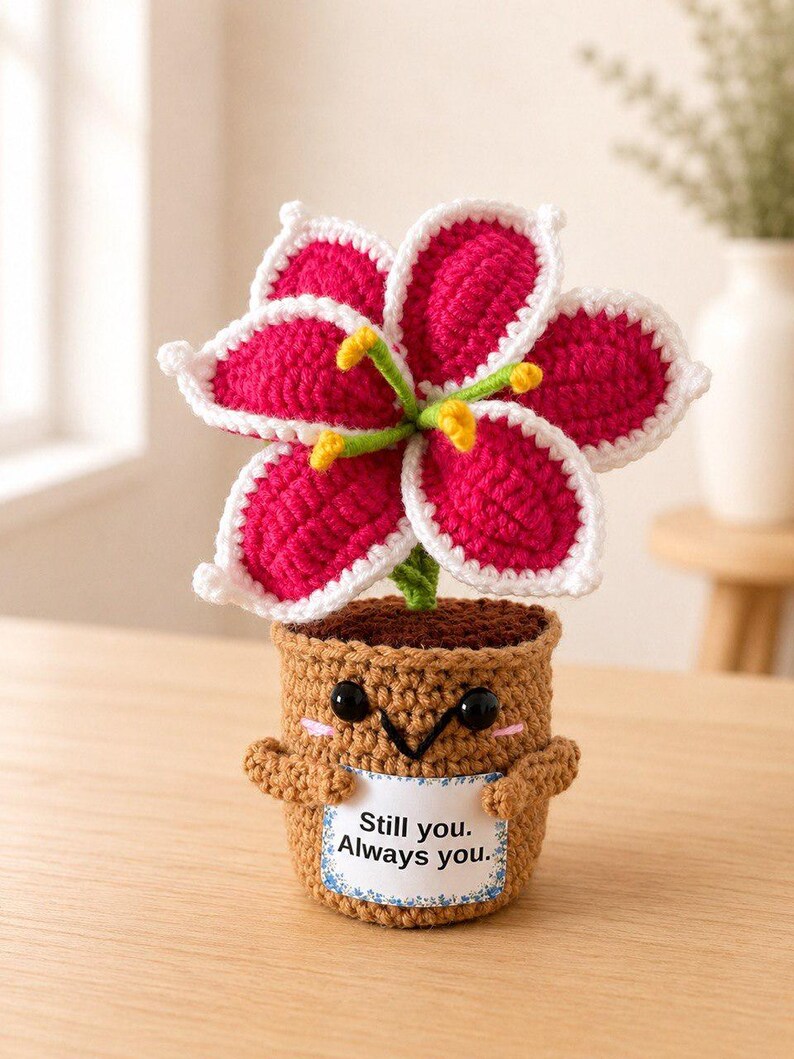 May include: A crocheted potted flower with vibrant magenta petals outlined in white, featuring yellow stamens. The brown crocheted pot has a smiling face, small arms, and a white sign that reads Still you. Always you. in black text.