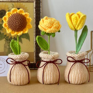 Könnte beinhalten: Drei handgefertigte Häkelblumen: eine Sonnenblume, eine Rose und eine Tulpe. Jede Blume ist gelb mit grünen Blättern und Stielen, in beige gestrickten Töpfen mit braunen Schleifen. Der Hintergrund zeigt ein gerahmtes Sonnenblumenbild.