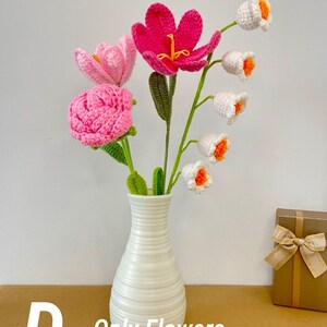 May include: A white ceramic vase holds a bouquet of handmade crochet flowers. The bouquet includes pink, fuchsia, and white flowers with orange centers. The vase sits on a brown surface. Text on the image reads "D - Only Flowers".