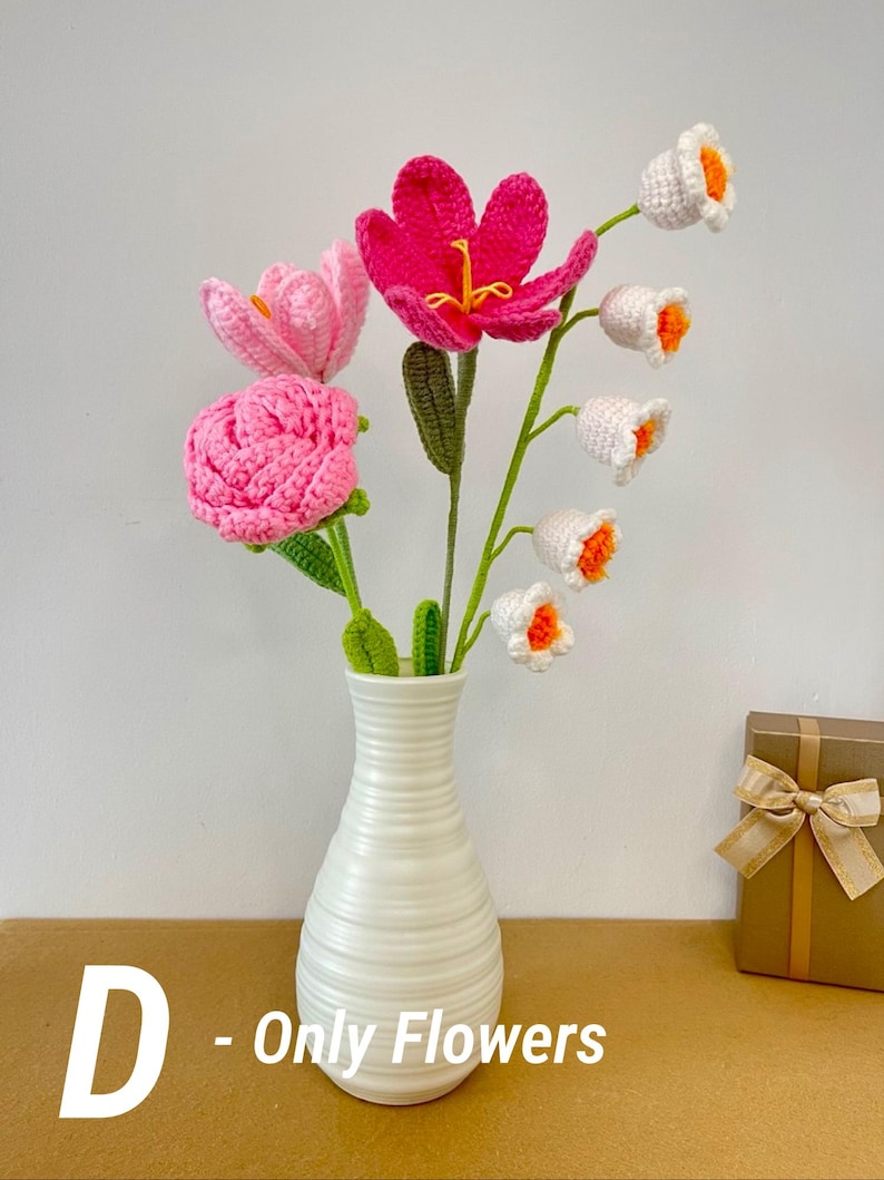 May include: A white ceramic vase holds a bouquet of handmade crochet flowers. The bouquet includes pink, fuchsia, and white flowers with orange centers. The vase sits on a brown surface. Text on the image reads "D - Only Flowers".